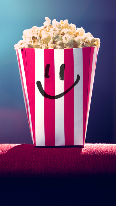 Ein gefüllter Popcorn-Becher mit Smiley-Gesicht vor einem leeren Kinosaal. Im Hintergrund blendet das Licht des Filmprojektors.