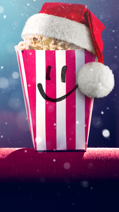Ein gefüllter Popcorn-Becher mit Smiley-Gesicht vor einem leeren Kinosaal. Im Hintergrund blendet das Licht des Filmprojektors.