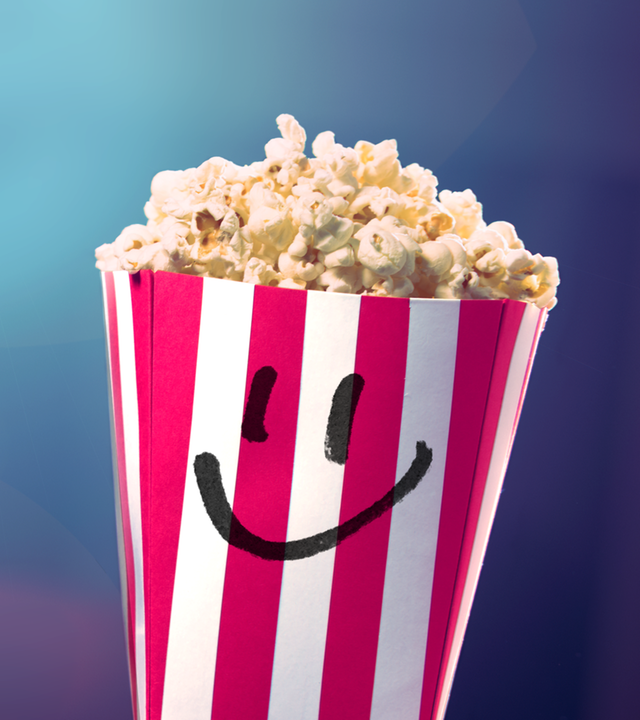 Ein gefüllter Popcorn-Becher mit Smiley-Gesicht vor einem leeren Kinosaal. Im Hintergrund blendet das Licht des Filmprojektors.
