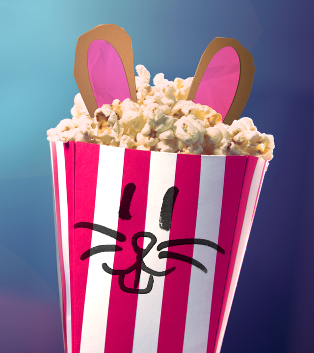 Ein gefüllter Popcorn-Becher mit Smiley-Hasen-Gesicht und Hasen-Ohren vor einem leeren Kinosaal. Im Hintergrund blendet das Licht des Filmprojektors.