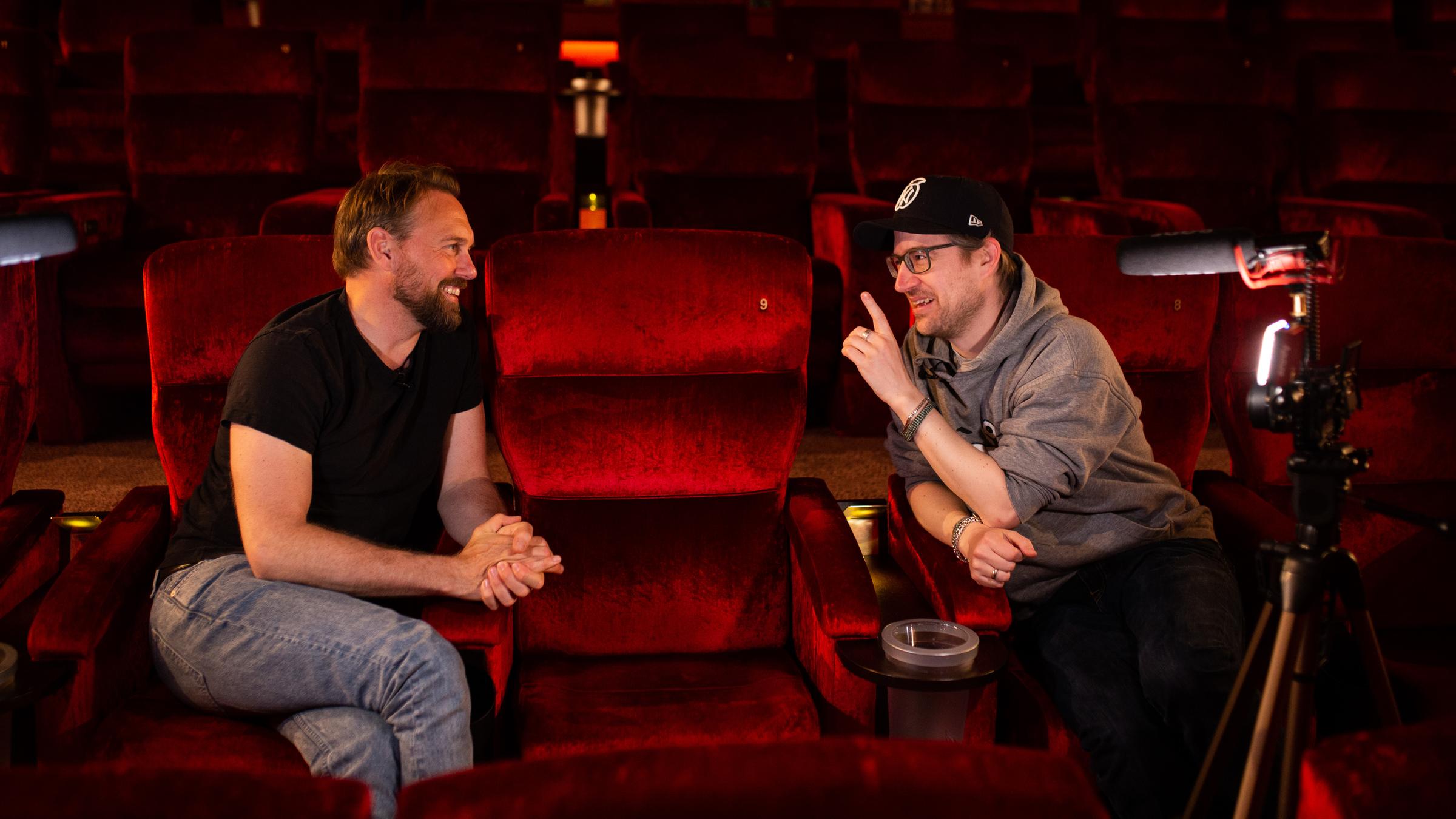 Steven Gätjen und Daniel Schröckert sitzen in einem Heimkino, schauen sich gegenseitig an und diskutieren miteinander.