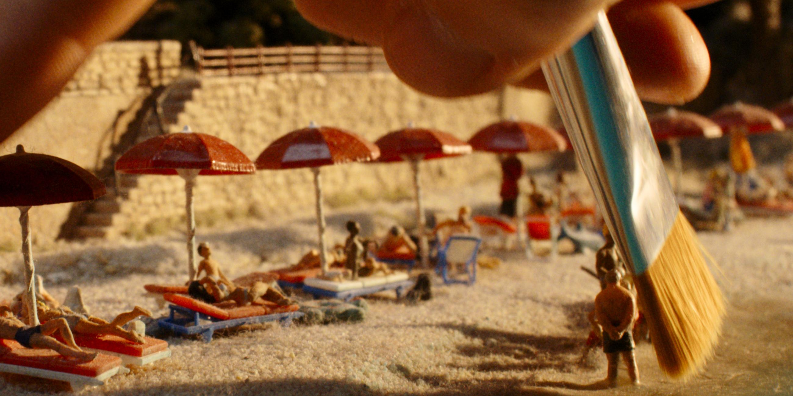 Eine Hand geht mit einem Pinsel über einen Miniaturstrand. An dem Stand liegen kleine Figürchen auf Strandliegen unter Sonnenschirmen.