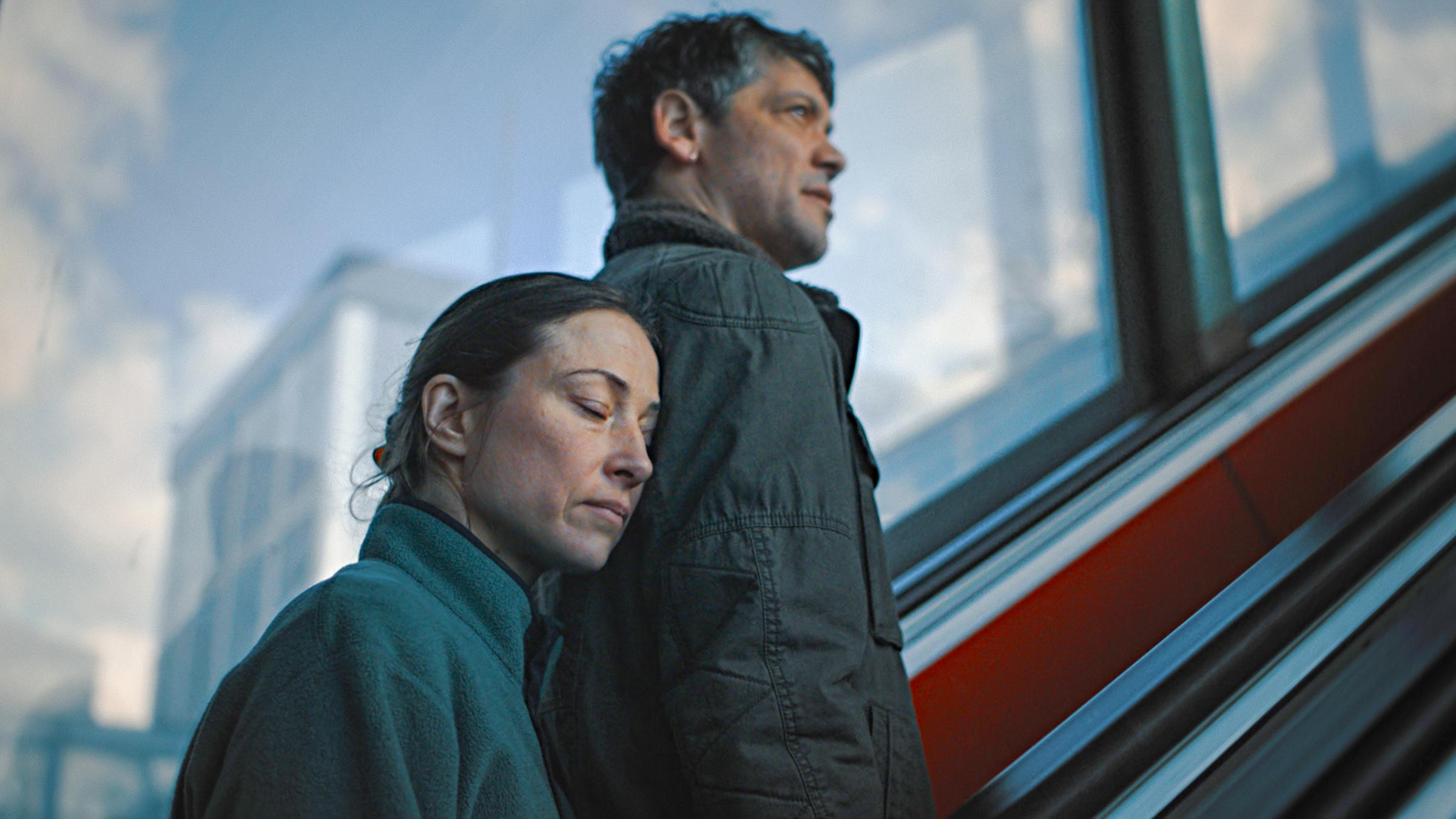 Eine Frau steht hinter einem Mann auf einer Rolltreppe und lehnt ihren Kopf mit geschlossenen Augen an ihn.