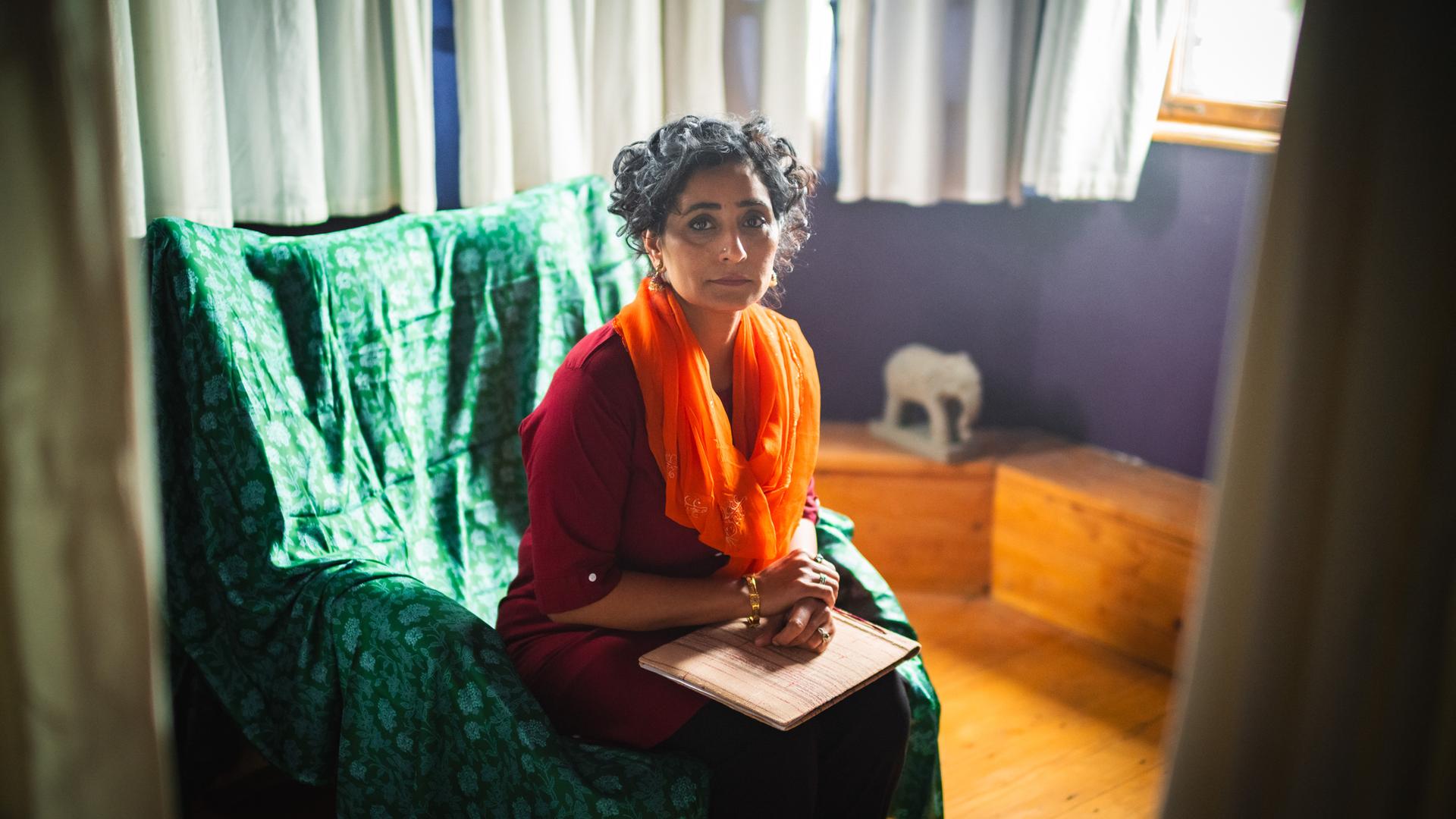 Szene aus "Meine Zuflucht und mein Sturm" von Arundhati Roy: Eine Fau sitzt auf einem grünen Sessel in einem Raum mit lila Wänden und weißen Vorhängen. Sie trägt ein rotes Oberteil und einen orangefarbenen Schal. Auf ihren Knien liegt ein Fotoalbum. Im Hintergrund steht eine kleine weiße Elefantenfigur auf einer Holzbank. Tageslicht fällt durch ein Fenster auf der rechten Seite ins Zimmer.