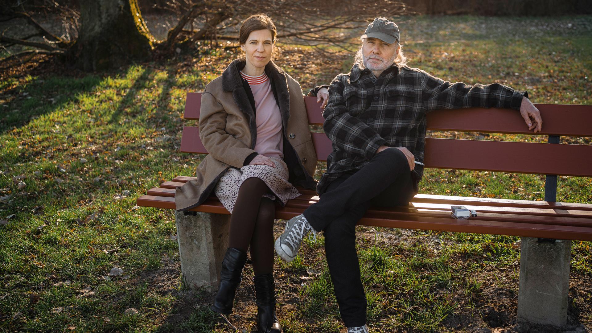 Nachgestellte Szene aus "Erzähl mir alles" von Elizabeth Strout: Eine Frau und ein Mann sitzen nebeneinander auf einer Holzbank in einem Park. Die Frau trägt einen hellen Mantel, ein Kleid mit schwarzen Stiefeln und sitzt mit übereinandergeschlagenen Beinen, während der Mann entspannt neben ihr sitzt und einen Arm auf der Rückenlehne der Bank ausstreckt. Er trägt eine karierte Jacke, dunkle Hose, Turnschuhe und eine Kappe. Neben ihm liegt eine Zigarettenschachtel auf der Bank. Im Hintergrund Bäume und winterliches Gras im warmen Sonnenlicht.
