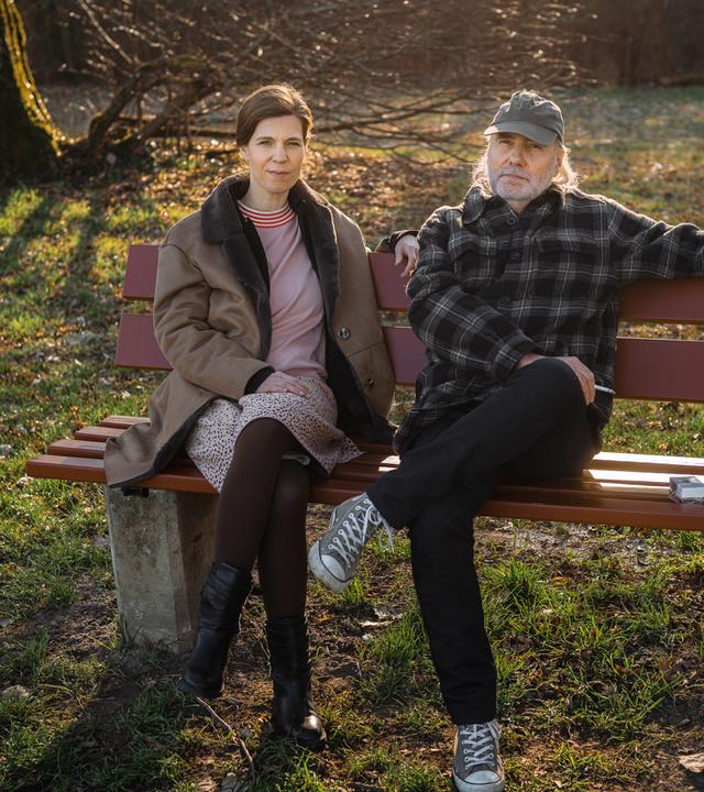 Nachgestellte Szene aus "Erzähl mir alles" von Elizabeth Strout: Eine Frau und ein Mann sitzen nebeneinander auf einer Holzbank in einem Park. Die Frau trägt einen hellen Mantel, ein Kleid mit schwarzen Stiefeln und sitzt mit übereinandergeschlagenen Beinen, während der Mann entspannt neben ihr sitzt und einen Arm auf der Rückenlehne der Bank ausstreckt. Er trägt eine karierte Jacke, dunkle Hose, Turnschuhe und eine Kappe. Neben ihm liegt eine Zigarettenschachtel auf der Bank. Im Hintergrund Bäume und winterliches Gras im warmen Sonnenlicht.