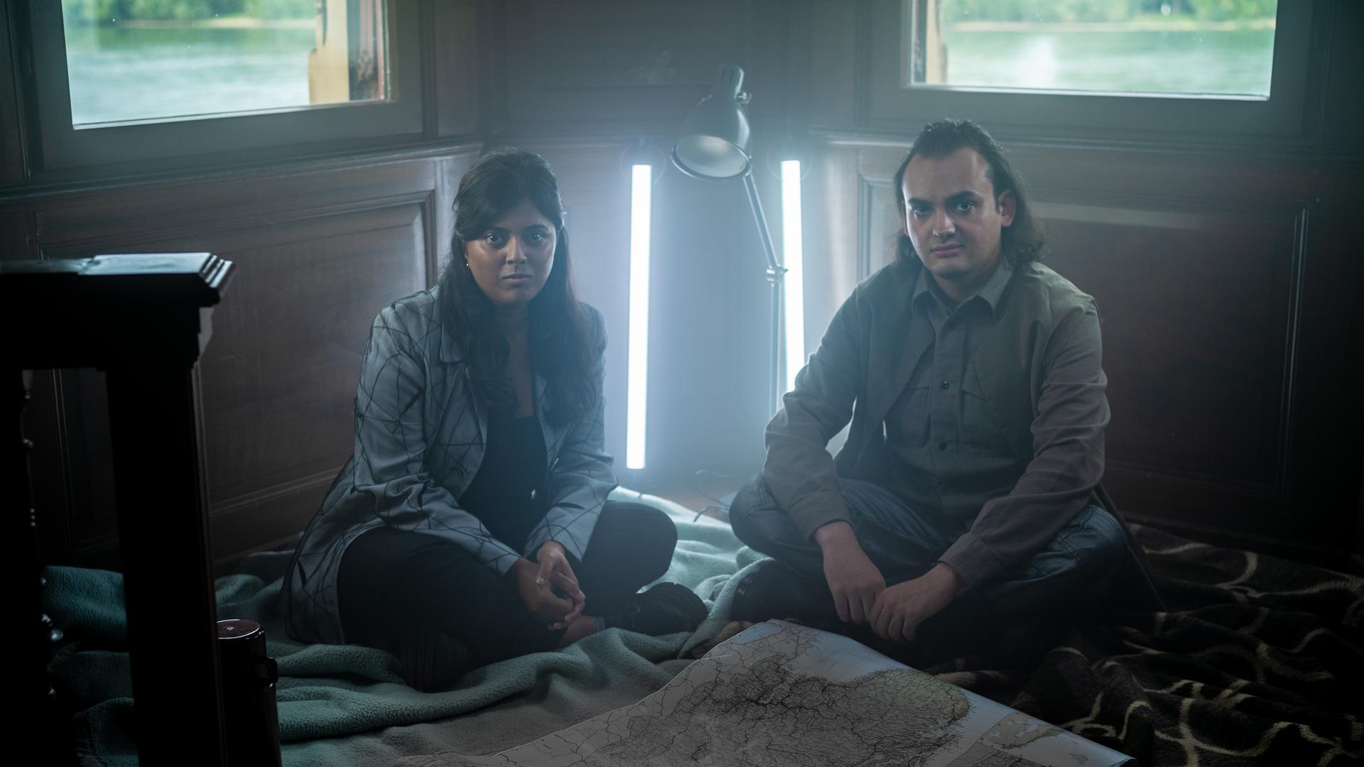 Szene aus "Was wir wissen können" von Ian McEwan: Zwei Personen sitzen auf dem Holzboden eines Zimmers mit zwei Fenstern, durch die man grüne Landschaft und Wasser sieht. Sie sitzen auf Decken, vor ihnen liegt eine große Landkarte ausgebreitet. Hinter ihnen stehen eine Lampe und zwei vertikale Leuchten, die die Szene beleuchten. Die Atmosphäre wirkt ruhig und konzentriert.