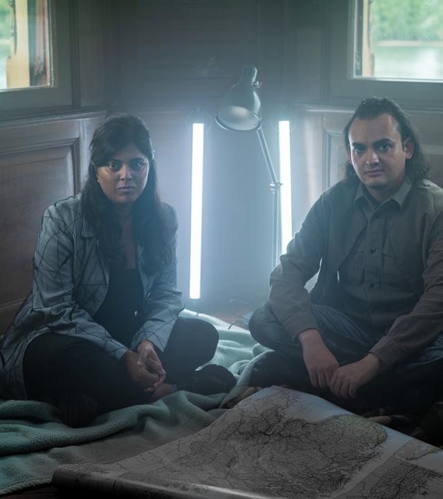 Szene aus "Was wir wissen können" von Ian McEwan: Zwei Personen sitzen auf dem Holzboden eines Zimmers mit zwei Fenstern, durch die man grüne Landschaft und Wasser sieht. Sie sitzen auf Decken, vor ihnen liegt eine große Landkarte ausgebreitet. Hinter ihnen stehen eine Lampe und zwei vertikale Leuchten, die die Szene beleuchten. Die Atmosphäre wirkt ruhig und konzentriert.