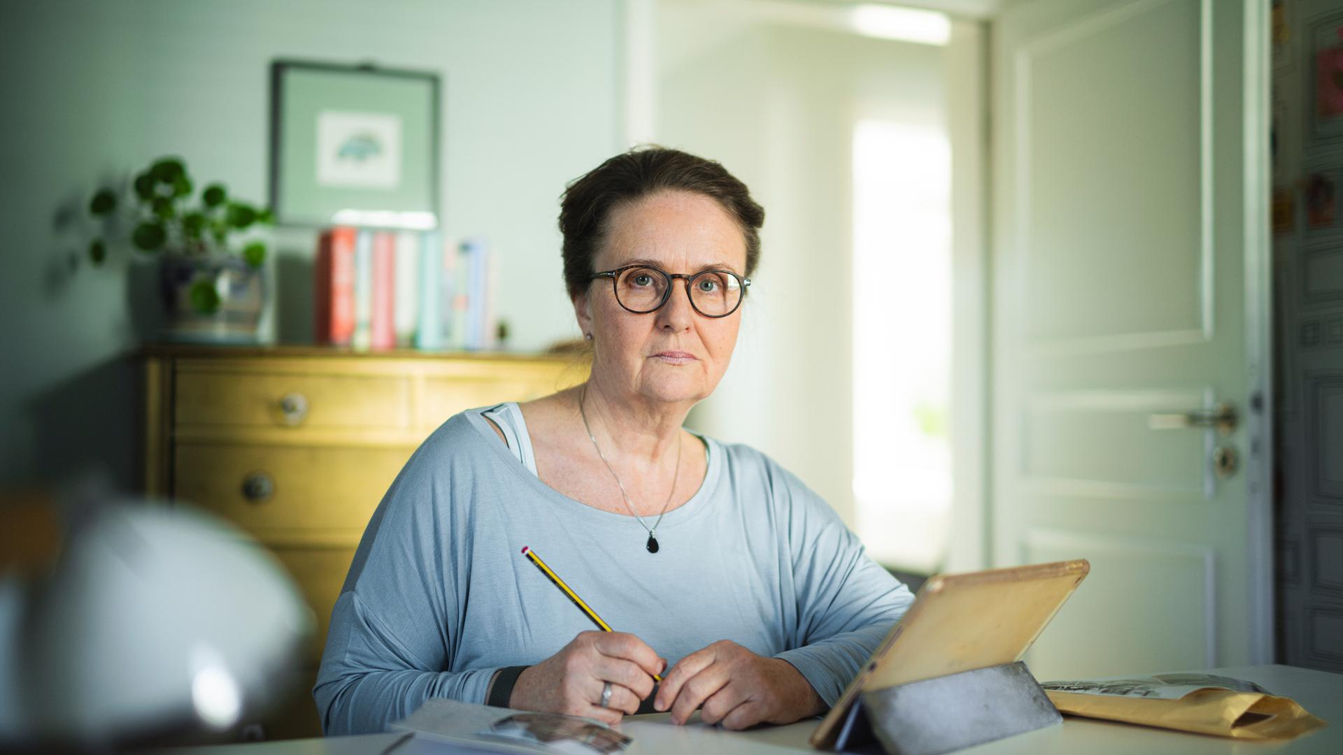 Nachgestellte Szene aus "Ich möchte zurückgehen in der Zeit" von Judith Hermann: Eine Frau sitzt an einem Tisch und hält einen Bleistift in der Hand. Sie trägt eine Brille und blickt mit ernstem Ausdruck nach vorne. Vor ihr stehen ein Tablet in einer Hülle, daneben liegt ein Notizbuch und ein brauner Briefumschlag. Hinter ihr befindet sich ein heller Raum mit einer Kommode, Büchern und einer Zimmerpflanze, durch eine offene Tür fällt Tageslicht hinein.