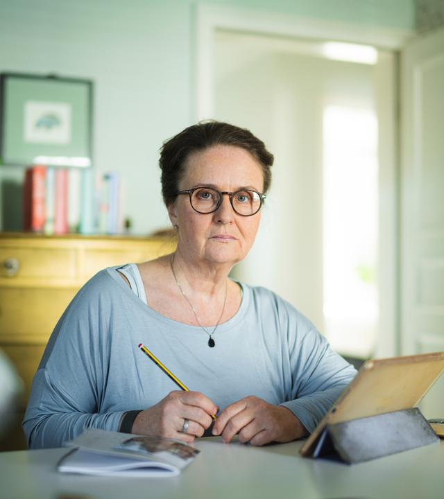 Nachgestellte Szene aus "Ich möchte zurückgehen in der Zeit" von Judith Hermann: Eine Frau sitzt an einem Tisch und hält einen Bleistift in der Hand. Sie trägt eine Brille und blickt mit ernstem Ausdruck nach vorne. Vor ihr stehen ein Tablet in einer Hülle, daneben liegt ein Notizbuch und ein brauner Briefumschlag. Hinter ihr befindet sich ein heller Raum mit einer Kommode, Büchern und einer Zimmerpflanze, durch eine offene Tür fällt Tageslicht hinein.