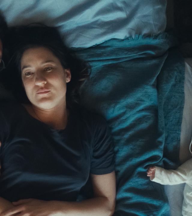 Ein Mann und eine Frau liegen auf ihrer Bettdecke im Bett. Der Mann hat die Augen geschlossen und lehnt seinen Kopf an den der Frau. Auch mit seinem Arm hält er sich an ihr fest. Die Frau hat die Augen geöffnet und blickt ins Leere. Neben ihnen im Beistellbett liegt ein schlafendes Baby.