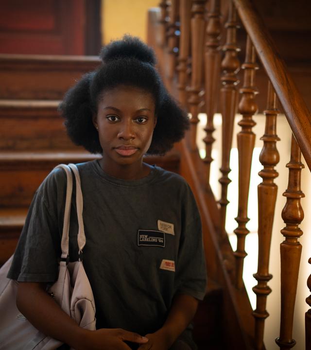 Szene aus "Der Fluch der Falodun Frauen" von Oyinkan Braithwaite: Eine junge Frau steht auf einer Holztreppe mit kunstvoll geschnitztem Geländer und blickt direkt in die Kamera. Sie trägt ein graues T-Shirt mit mehreren Aufnähern und hat eine helle Tasche über der Schulter. Im Hintergrund befindet sich eine rote Tür.