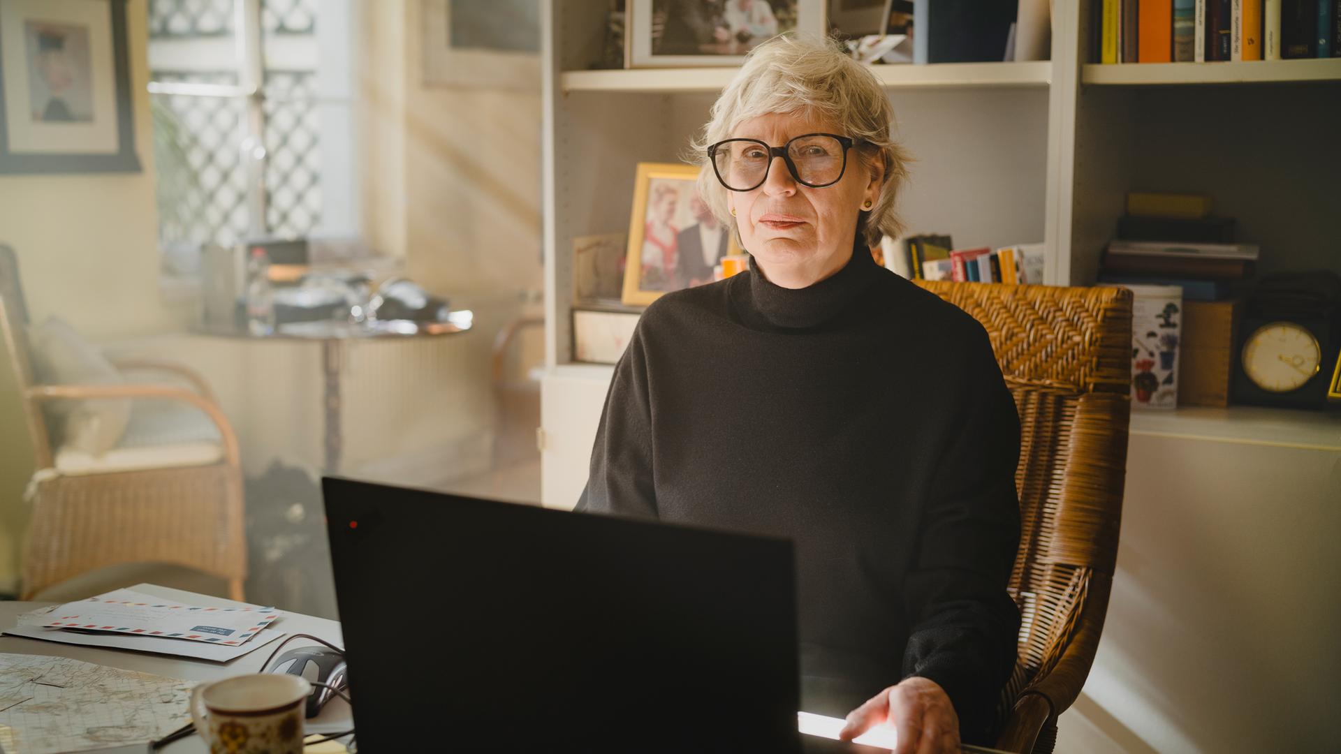 Nachgestellte Szene aus "Ghost Stories. Ein Buch der Erinnerung" von Siri Hustvedt: Eine ältere Frau mit kurzen grauen Haaren und runder Brille sitzt in einem Korbstuhl vor einem Laptop. Sie trägt einen schwarzen Rollkragenpullover. Vor ihr stehen eine Tasse und mehrere Briefe, im Hintergrund sind Bücherregale, Familienfotos und eine Uhr zu sehen. Durch ein Fenster fällt warmes Tageslicht in den Raum und erzeugt eine ruhige Atmosphäre.