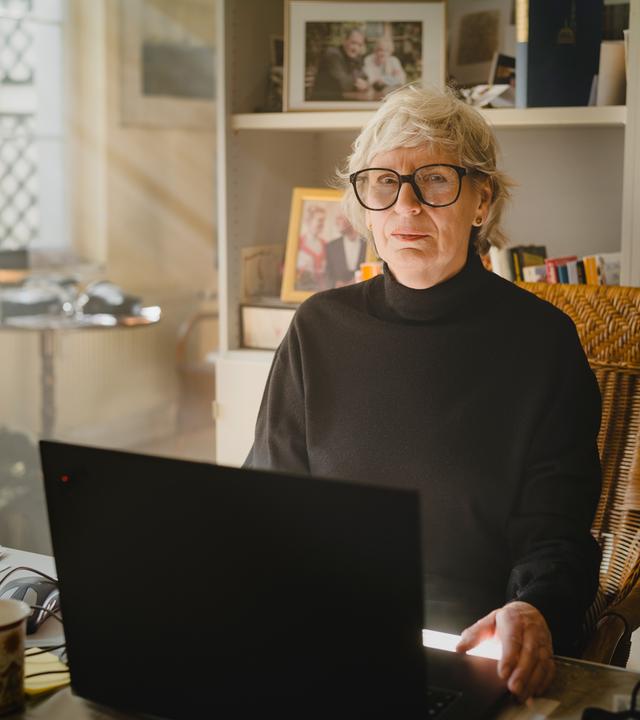 Nachgestellte Szene aus "Ghost Stories. Ein Buch der Erinnerung" von Siri Hustvedt: Eine ältere Frau mit kurzen grauen Haaren und runder Brille sitzt in einem Korbstuhl vor einem Laptop. Sie trägt einen schwarzen Rollkragenpullover. Vor ihr stehen eine Tasse und mehrere Briefe, im Hintergrund sind Bücherregale, Familienfotos und eine Uhr zu sehen. Durch ein Fenster fällt warmes Tageslicht in den Raum und erzeugt eine ruhige Atmosphäre.