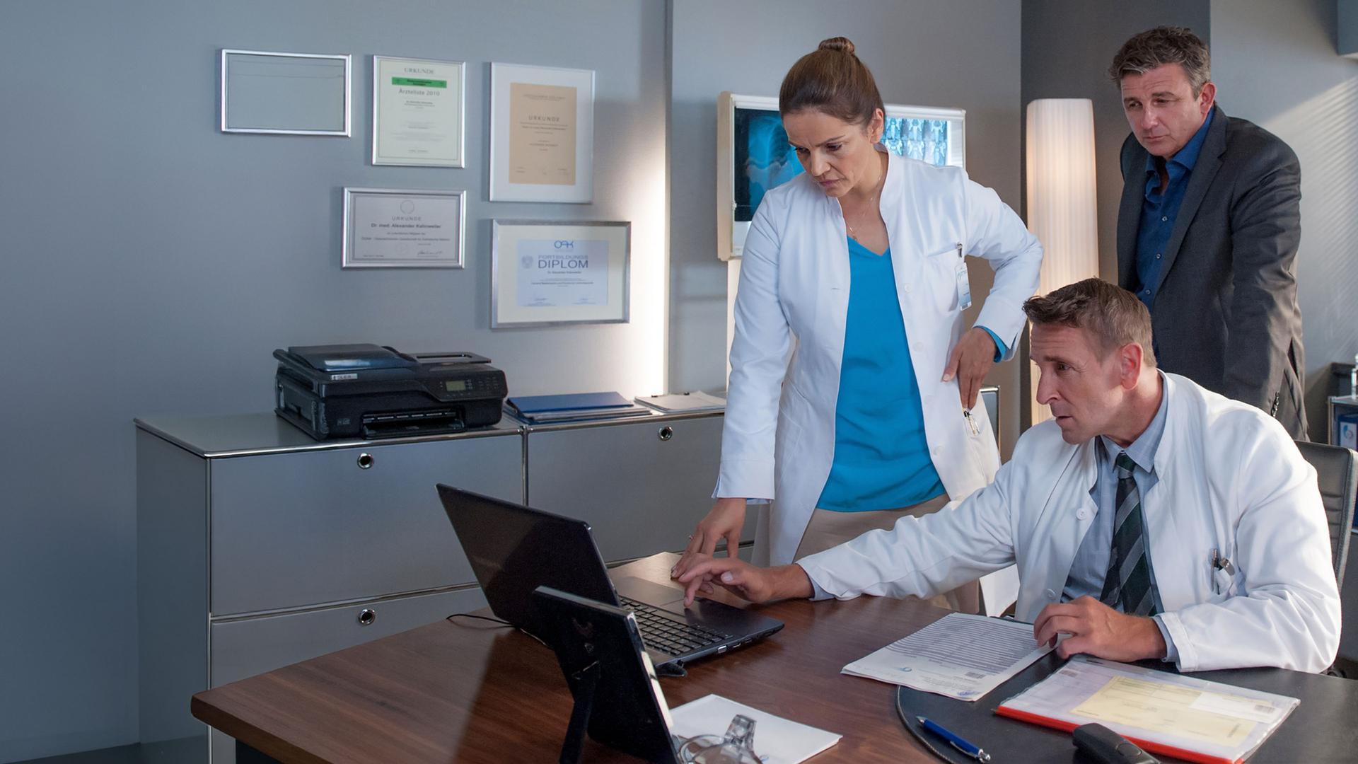 "Der Bergdoktor: Finale Klarheit (2)": In einem Büro. Vera (Rebecca Immanuel), Alexander (Mark Keller) und Martin (Hans Sigl) blicken ungläubig auf einen auf dem Tisch stehenden Monitor.