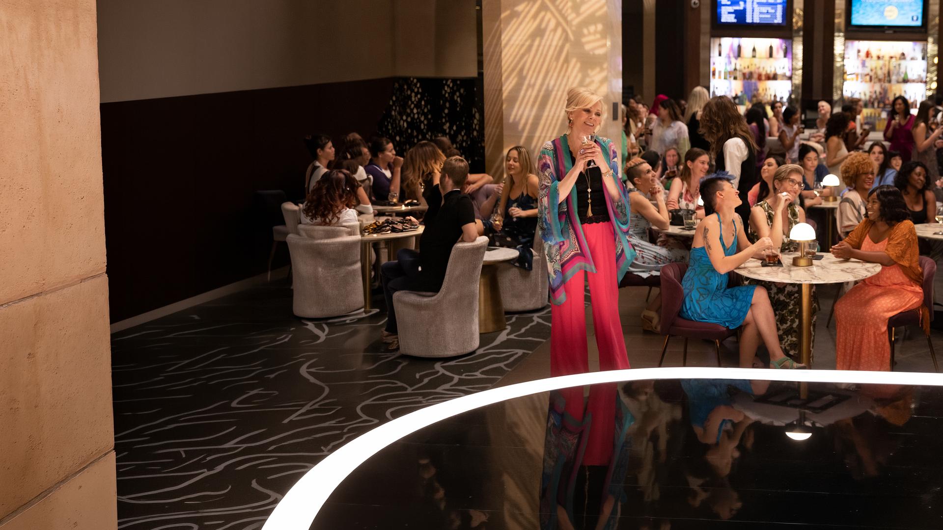 "Hacks - Findet mich toll, einfach": Deborah (Jean Smart) steht lächelnd mit einem Martini-Glas in der Hand, in einer gut besuchten Bar vor einer Bühne, während um sie herum weibliche Gäste an Tischen sitzen und sich unterhalten.