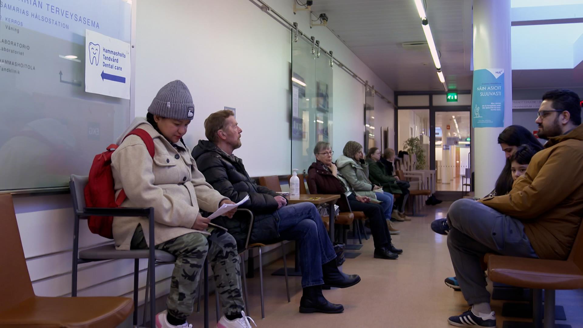 Menschen sitzen in einem gefüllten Warteraum, vermutlich in einem Krankenhaus