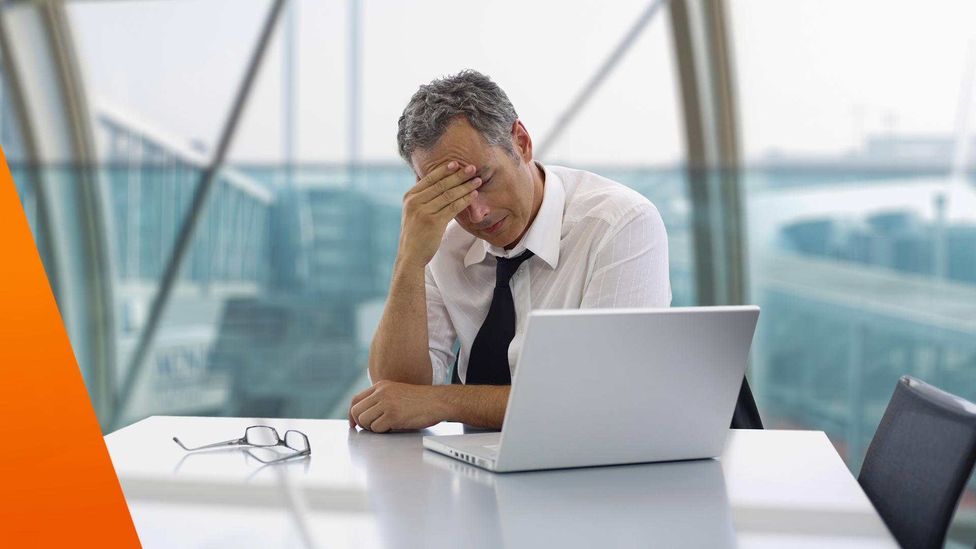 Ein Mann sitzt in einem modernen Büro an einem Tisch mit Laptop und wirkt nachdenklich und belastet. Er hat die Augen geschlossen oder gesenkt und stützt den Kopf mit der Hand. Auf dem Tisch liegt eine Brille, im Hintergrund sind große Fenster mit Blick nach draußen.