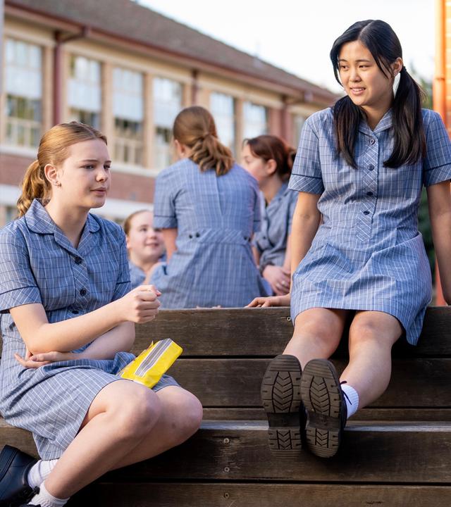 Hannah, Olivia und Natalie sitzen auf den Holzstufen draußen auf dem Pausenhof und unterhalten sich. Hannah schaut nachdenklich aus, während Olivia und Natalie ihr zuhören. Im Hintergrund sitzen weitere Schülerinnen.