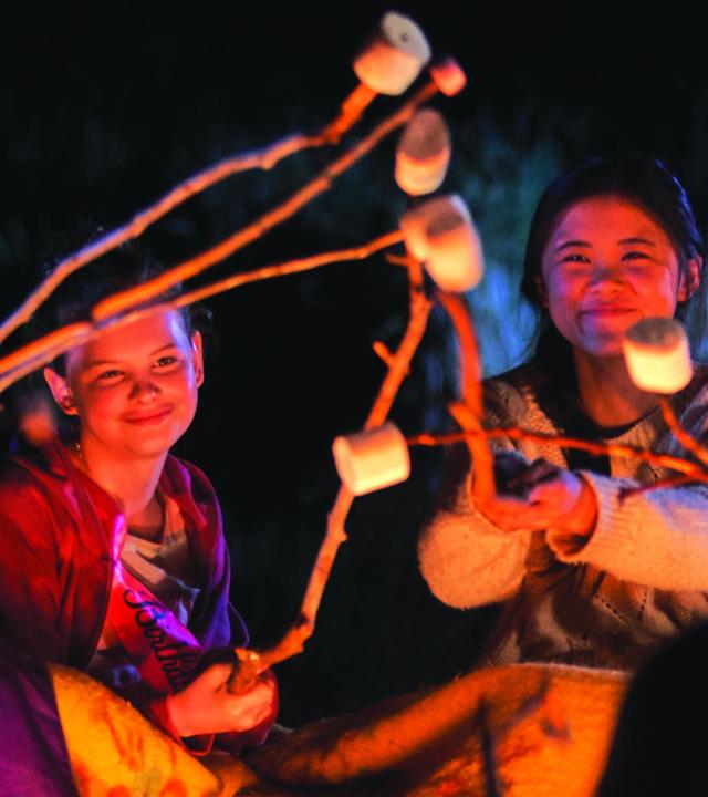 Natalie, Hannah, Olivia, Sarah und andere Mitschülerinnen sitzen abends um ein Lagerfeuer herum. In ihren Händen halten sie Stöcke auf denen Marshmallows aufgespießt sind. Die Stimmung ist ausgelassen und alle Mädchen lachen.