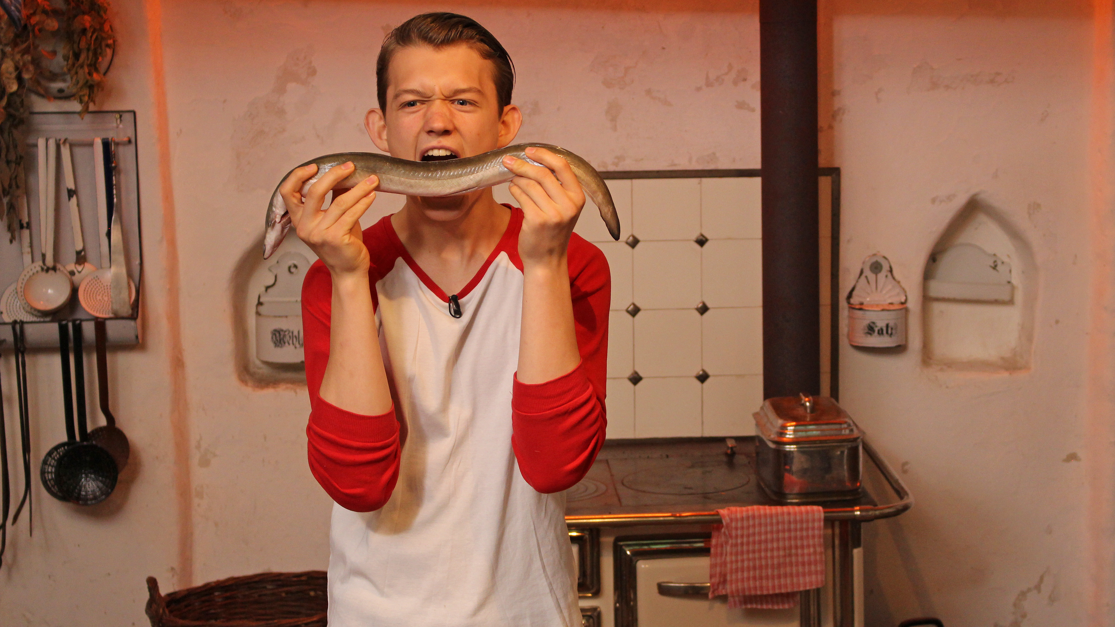 EIn Junge steht in einer Küche und hat einen Aal in der Hand. Er tut so, als würde er hineinbeißen.