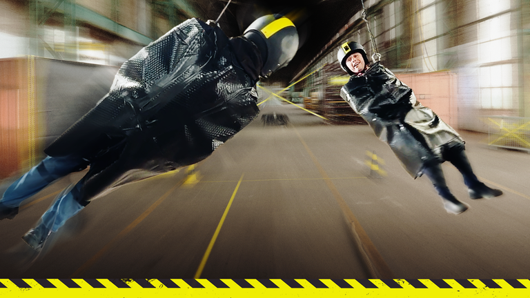 Checkpoint: Die Testteamer Cem und Mark sind in feste Plastikmäntel eingerollt, tragen Schutzhelme und hängen wie Pendel von einem Stahlseil, das in einem Klettergurt befestigt ist. 