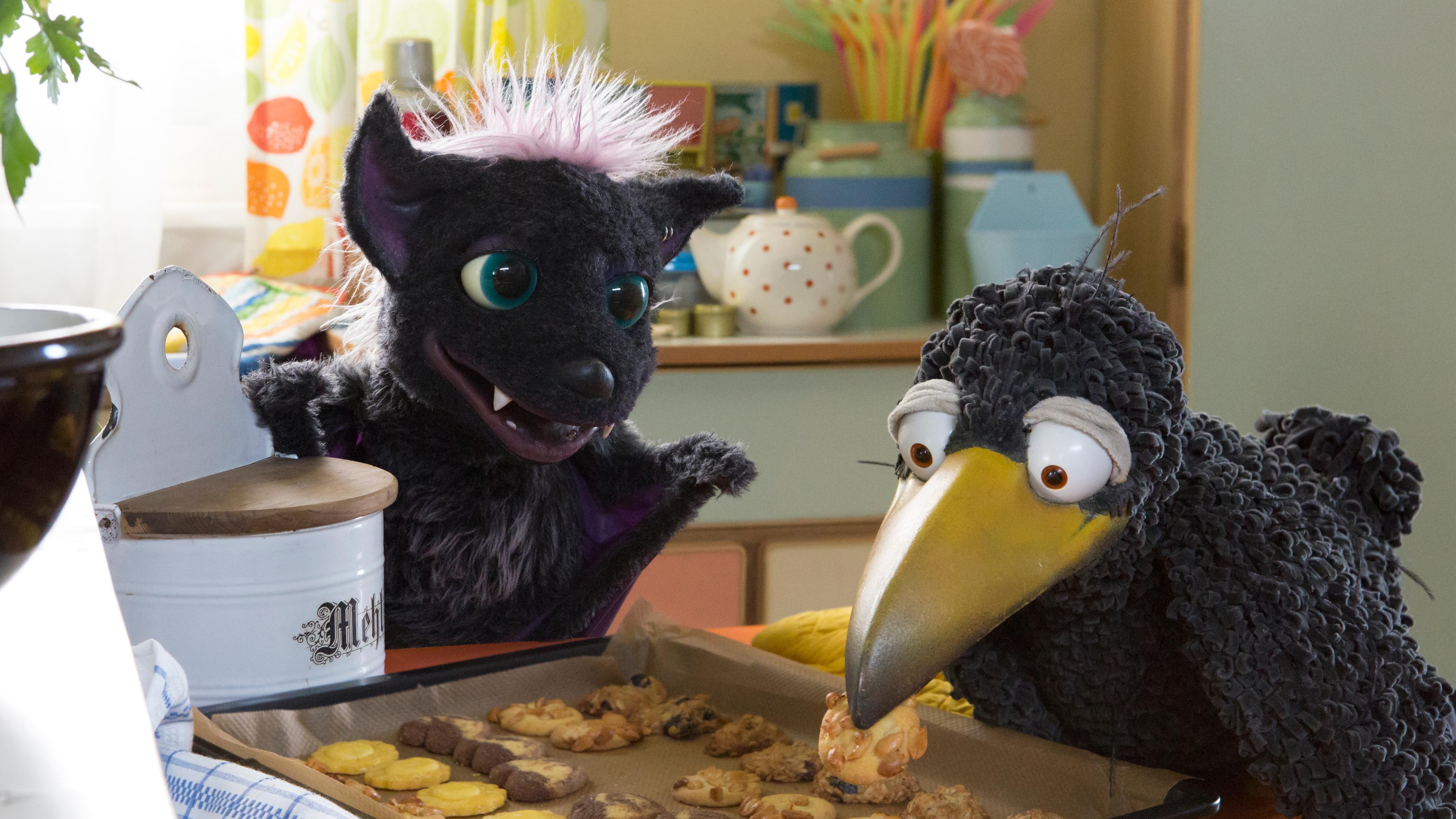 Rabe Rudi und Vampi hocken auf dem Küchentisch und schauen begierig auf ein Blech voller Kekse.