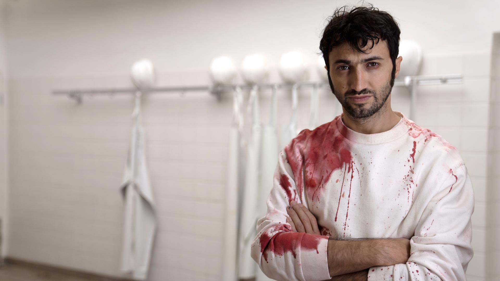 "Fleisch":  Adrian (Axel Moustache) steht in einem weiß gekachelten Raum und blickt in die Kamera. Sein weißer Arbeitspullover ist blutverschmiert. Er hat die Hände vor dem Körper verschränkt.