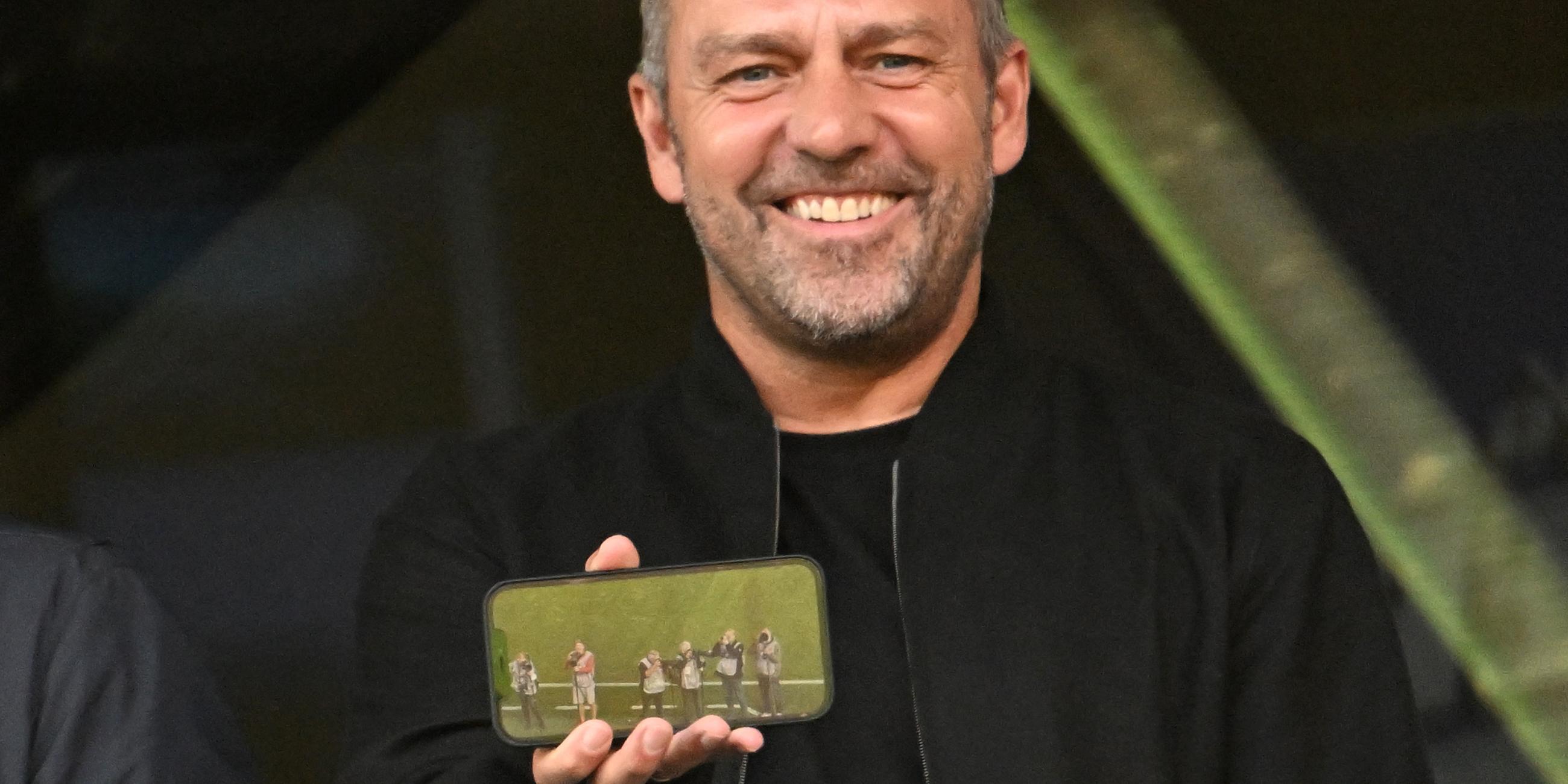 Fußball-Bundesliga, Bayer Leverkusen - VfL Wolfsburg: Bundestrainer Hansi Flick sitzt auf der Tribüne und zeigt den Fotografen ein Bild auf seinem Smartphone.