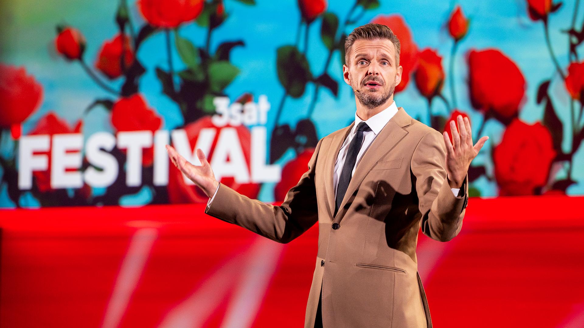 Kabarettist steht in der Mitte des Bildes und breitet seine Arme seitlich aus. Die Bühne ist rot beleuchtet und es sind Rosen auf der Vidiwall abgebildet. Florian Schroeder trägt einen beigen Anzug.