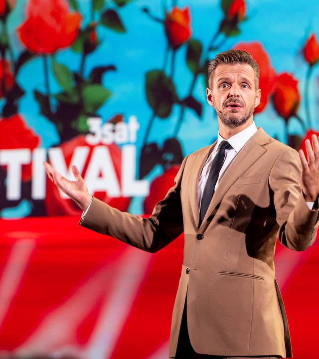 Kabarettist steht in der Mitte des Bildes und breitet seine Arme seitlich aus. Die Bühne ist rot beleuchtet und es sind Rosen auf der Vidiwall abgebildet. Florian Schroeder trägt einen beigen Anzug.