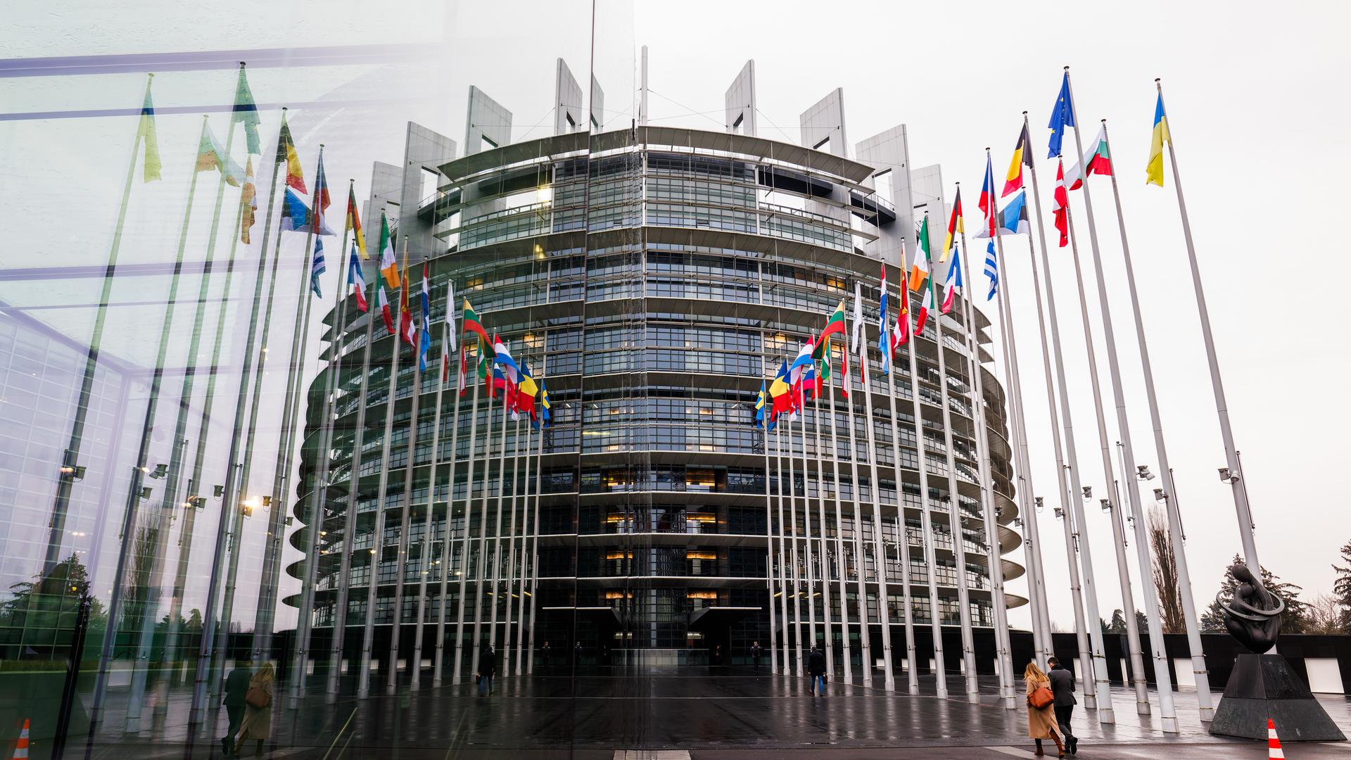 Die Fahnen der Mitgliedsstaaten der Europäischen Union und der Ukraine wehen vor dem Gebäude des Europäischen Parlaments und spiegeln sich dabei in einer gläsernen Wand.