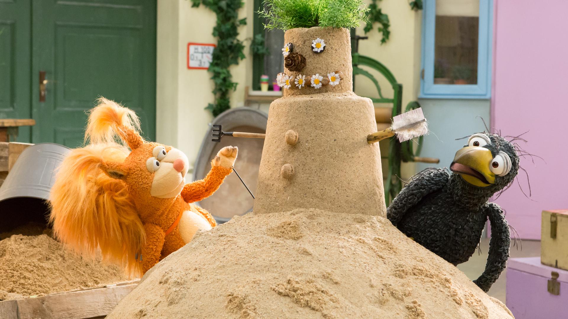 Ella und Rudi stehen rechts und links vor einem selbstgebauten Sandmann. Er hat Gänseblümchenaugen, eine Zapfennase, eine Grünpflanze als Haarschopf und eine Harke und eine Bürste als Arme.