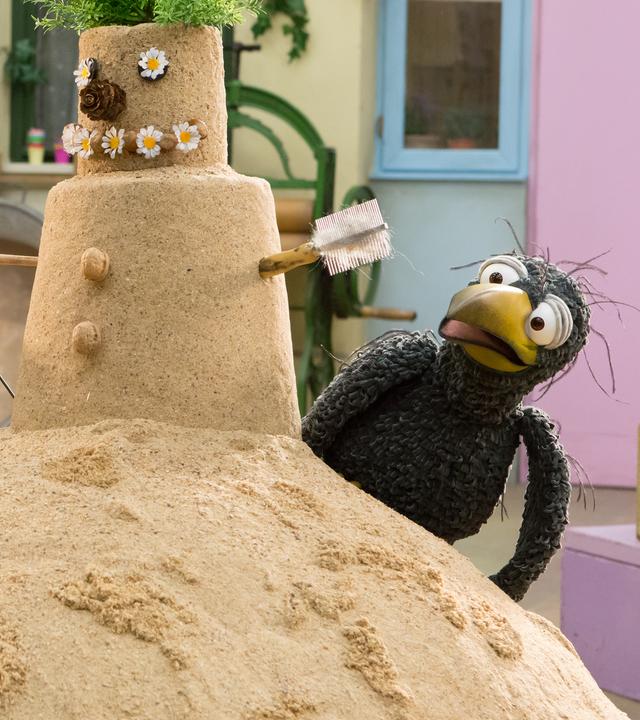 Ella und Rudi stehen rechts und links vor einem selbstgebauten Sandmann. Er hat Gänseblümchenaugen, eine Zapfennase, eine Grünpflanze als Haarschopf und eine Harke und eine Bürste als Arme.