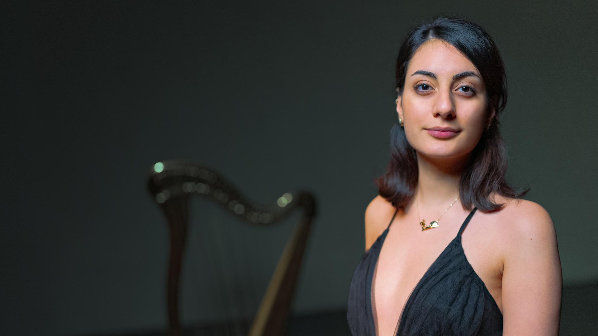 Toki trägt ein schwarzes Kleid und schaut leicht lächelnd frontal in die Kamera. Im Hintergrund der Schemen einer Harfe.