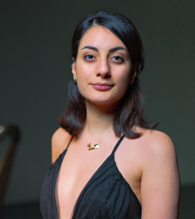 Toki trägt ein schwarzes Kleid und schaut leicht lächelnd frontal in die Kamera. Im Hintergrund der Schemen einer Harfe.