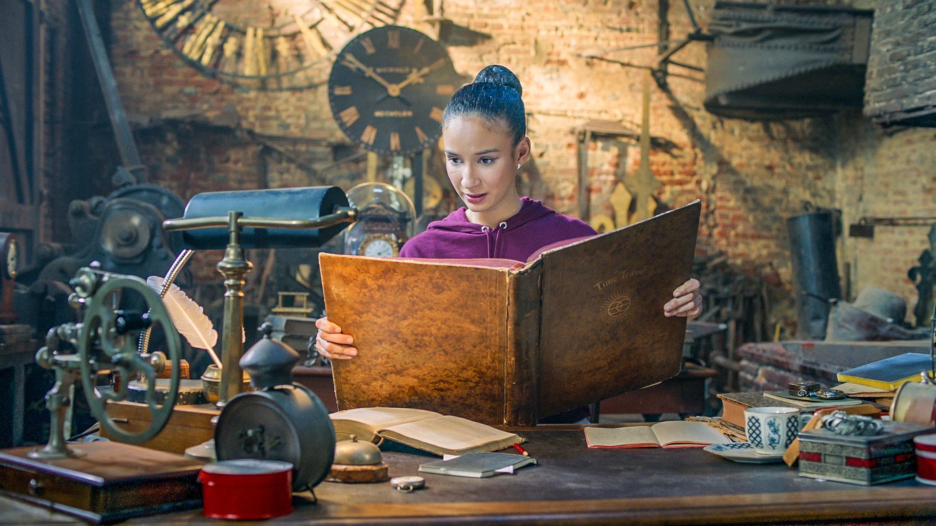 Ines steht in Oscars Werkstatt. Sie hält ein großes, braunes Buch in der Hand, in dem sie liest.