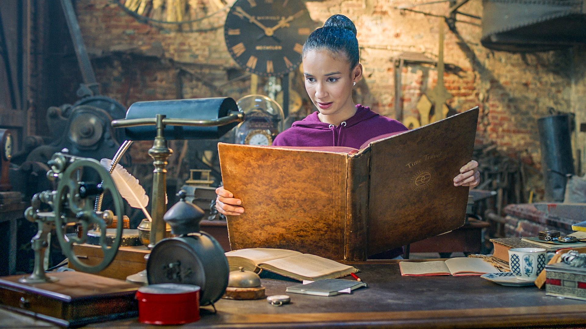 Ines steht in Oscars Werkstatt. Sie hält ein großes, braunes Buch in der Hand, in dem sie liest.
