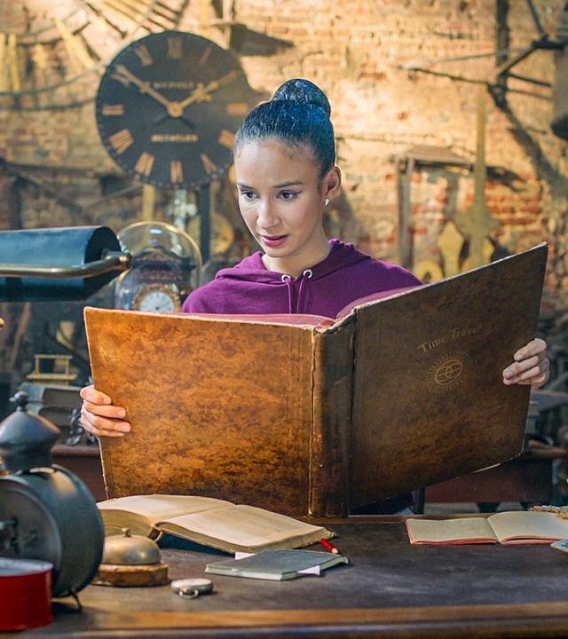 Ines steht in Oscars Werkstatt. Sie hält ein großes, braunes Buch in der Hand, in dem sie liest.