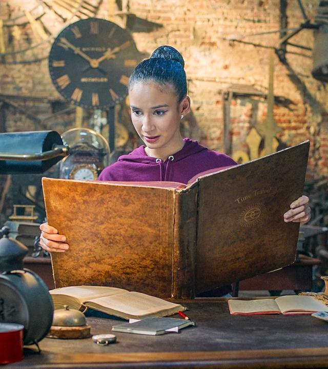 Ines steht in Oscars Werkstatt. Sie hält ein großes, braunes Buch in der Hand, in dem sie liest.