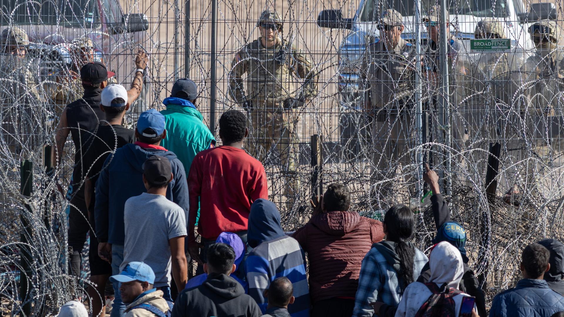 Rückansicht von Menschen dunklerer Hautfarbe, die vor einem Stacheldrahtzaun stehen. Auf der anderen Seite stehen Soldaten in Camouflage.