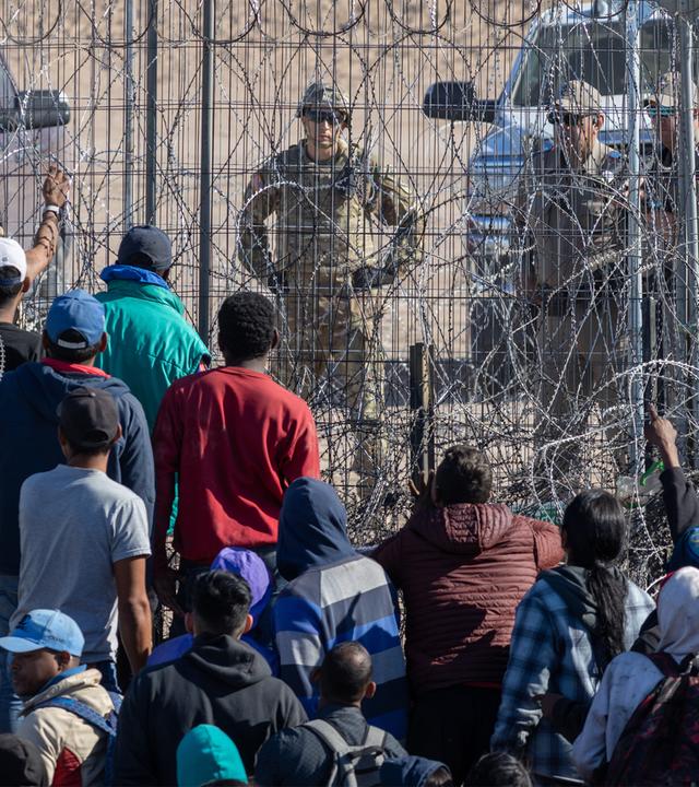 Rückansicht von Menschen dunklerer Hautfarbe, die vor einem Stacheldrahtzaun stehen. Auf der anderen Seite stehen Soldaten in Camouflage.