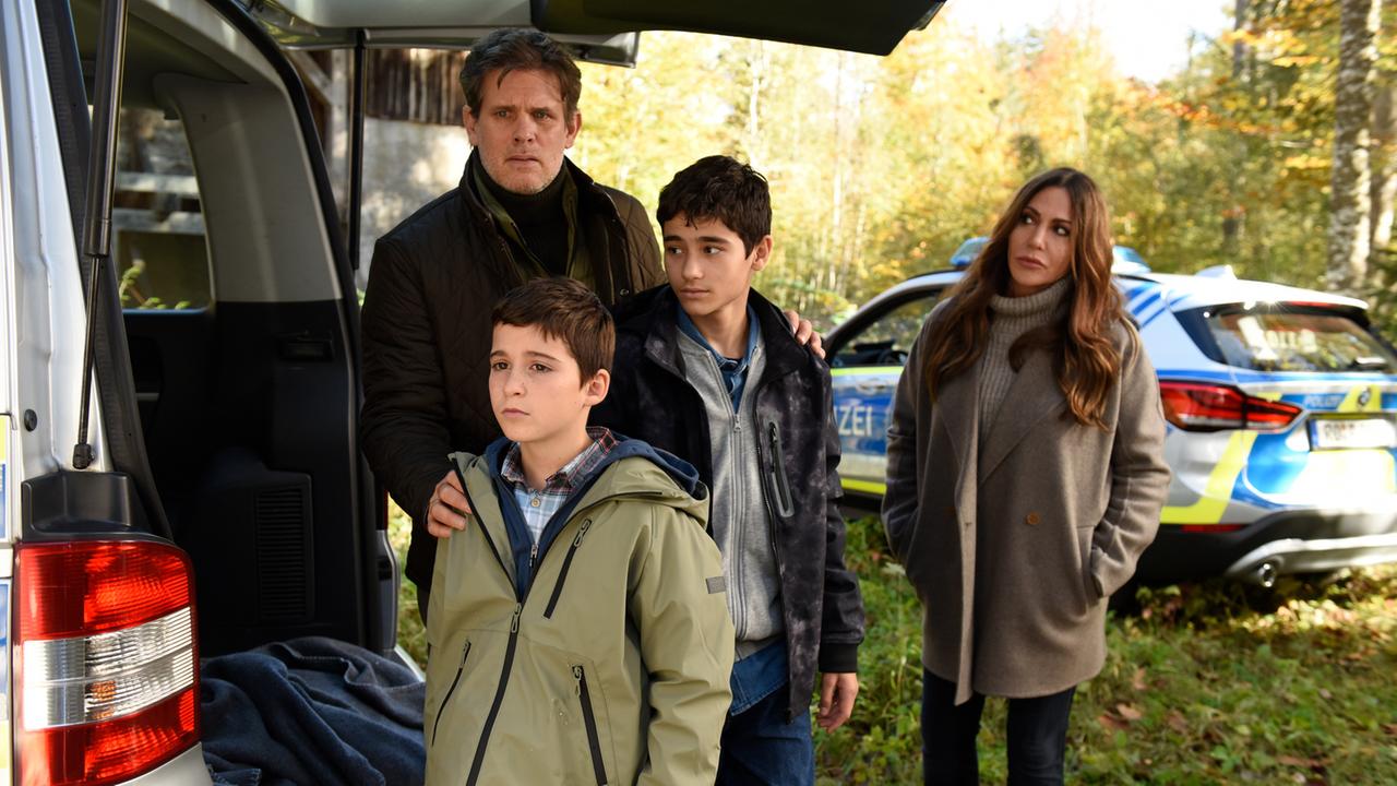 "Frühling: Flüsternde Geister": Alexander Jacobi (Steffen Groth) steht mit seinen Kindern Sammy (Vico-Simone Magno) und Milo (Mika Ullritz) neben einem Pkw. Er schaut besorgt. Katja Baumann (Simone Thomalla) steht abseits daneben und schaut ihn mit fürsorglichen Blick an. Im Hintergrund sieht man ein Polizeiauto.