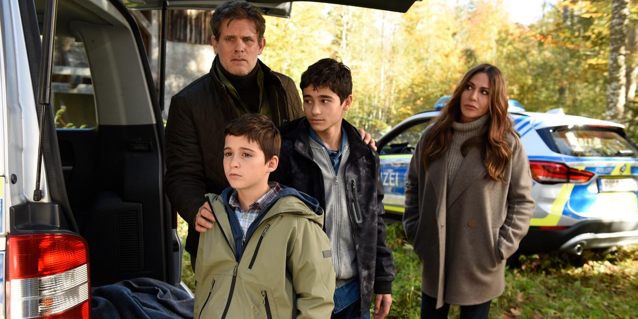 "Frühling: Flüsternde Geister": Alexander Jacobi (Steffen Groth) steht mit seinen Kindern Sammy (Vico-Simone Magno) und Milo (Mika Ullritz) neben einem Pkw. Er schaut besorgt. Katja Baumann (Simone Thomalla) steht abseits daneben und schaut ihn mit fürsorglichen Blick an. Im Hintergrund sieht man ein Polizeiauto.