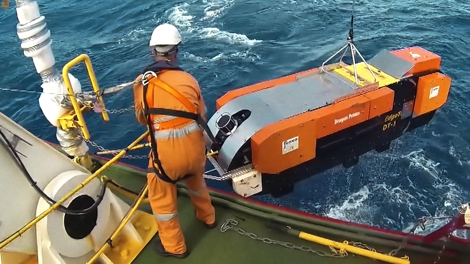 Ein Mann im orangenen Overall steht auf einer Plattform im Meer und lässt mithilfe einer Winde einen orangenen, etwa zwei Meter langen Tauchroboter zu Wasser.