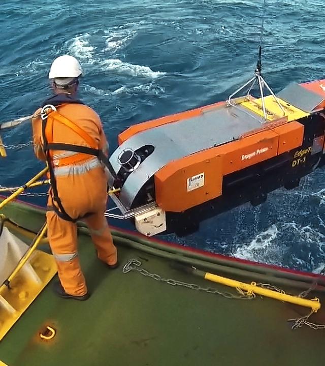 Ein Mann im orangenen Overall steht auf einer Plattform im Meer und lässt mithilfe einer Winde einen orangenen, etwa zwei Meter langen Tauchroboter zu Wasser.