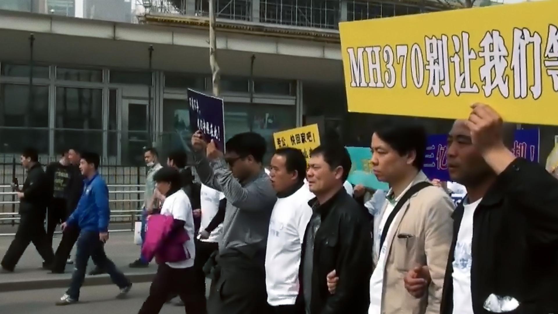 Eine Gruppe Menschen gehen in einer Reihe eine Straße entlang. Sie tragen ein Schild, auf dem in weißen Buchstaben auf gelbem Grund MH370 steht, daneben stehen chinesische Schriftzeichen.