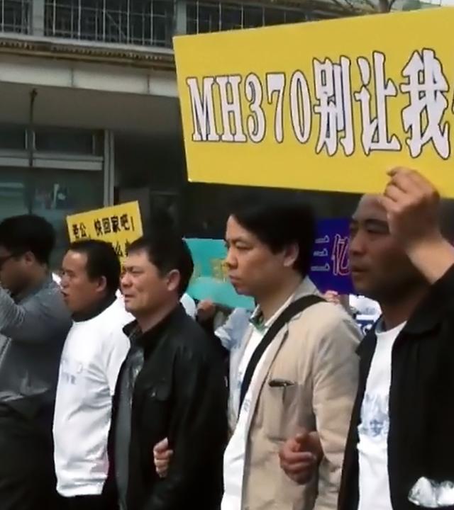 Eine Gruppe Menschen gehen in einer Reihe eine Straße entlang. Sie tragen ein Schild, auf dem in weißen Buchstaben auf gelbem Grund MH370 steht, daneben stehen chinesische Schriftzeichen.