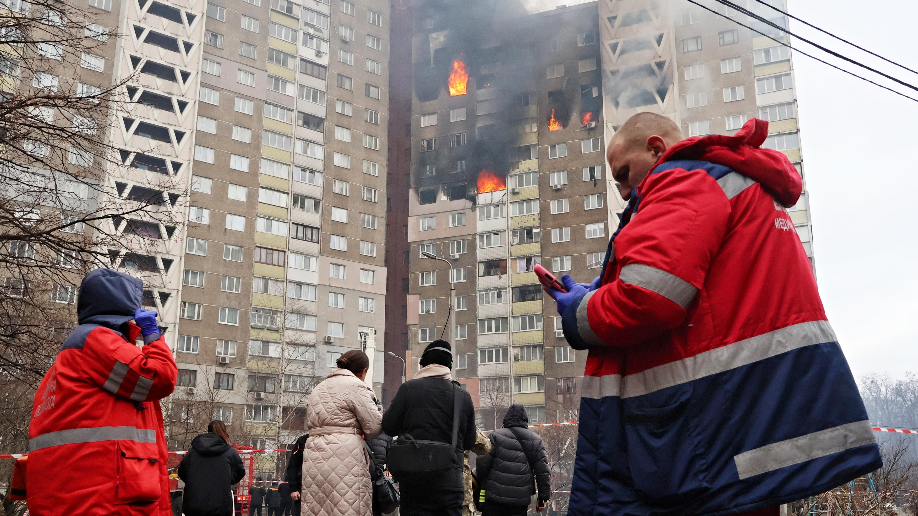 Rettungskräfte und Zivilisten stehen vor einem brennenden Wohngebäude, dass von russischen Marschflugkörpern getroffen wurde.