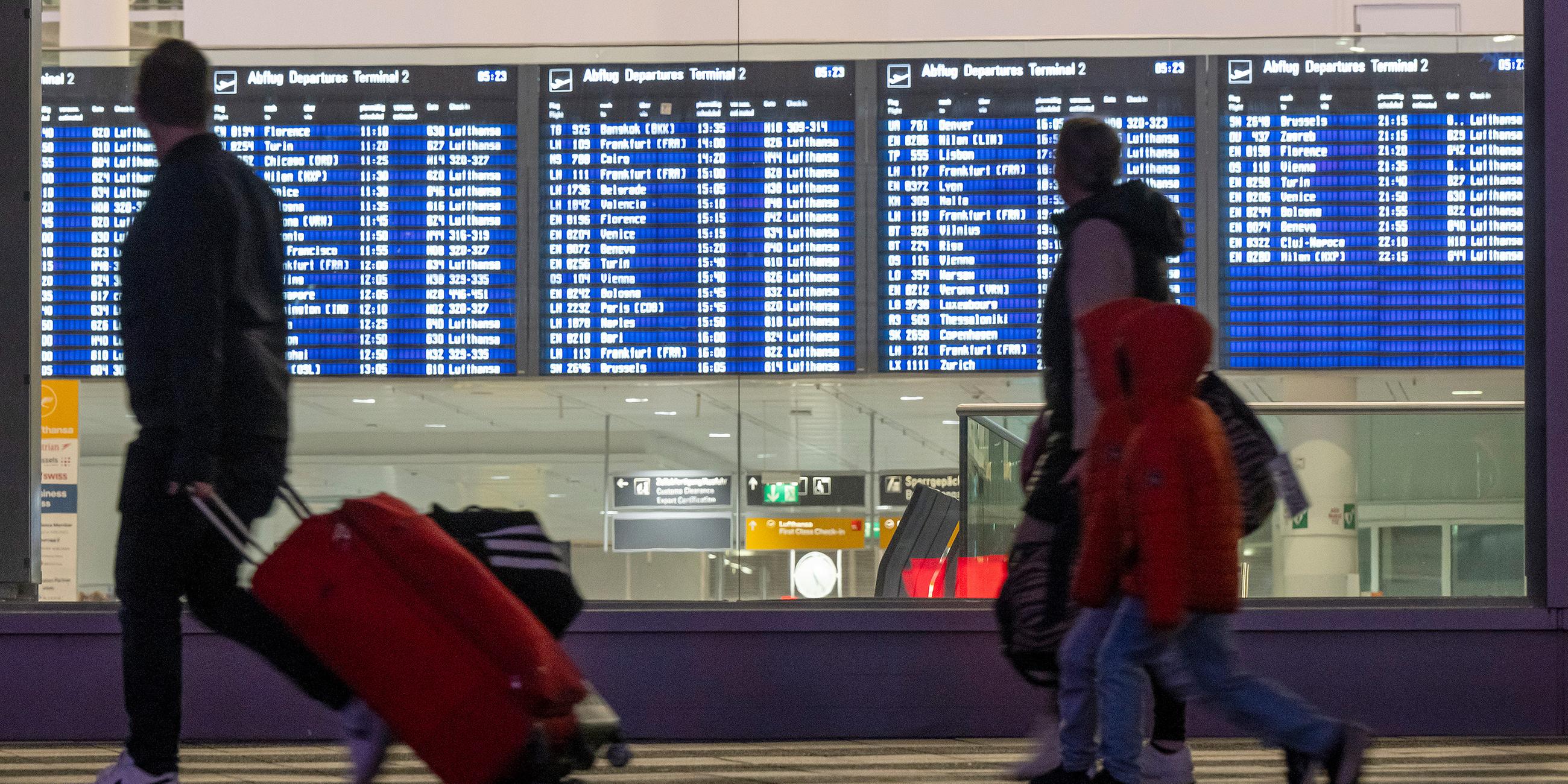 Eine Familie mit Koffern läuft an einer Anzeigetafel für die Abflüge am Flughafen München vorbei